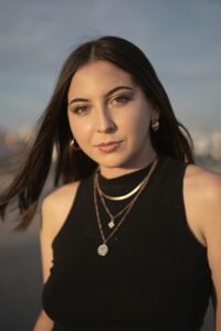Portrait of a young woman with jewelry, black crop top, and windblown hair at sunset.